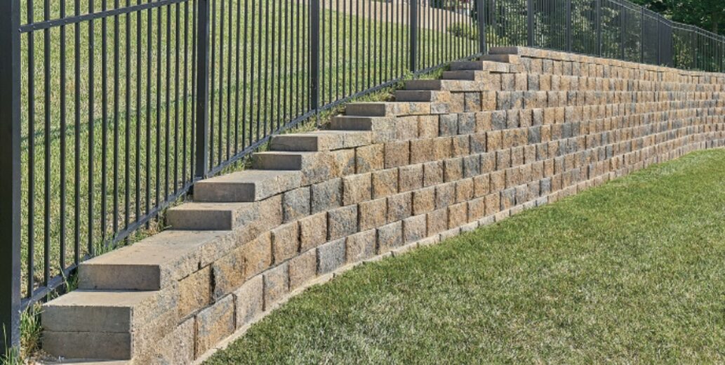 Stepped tan block retaining wall following a slope beside a metal fence