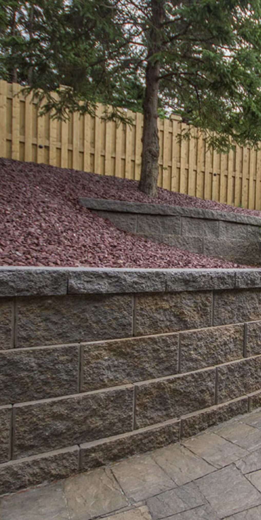 Tall terraced split-face block retaining wall with fresh gravel beds and a wood fence above